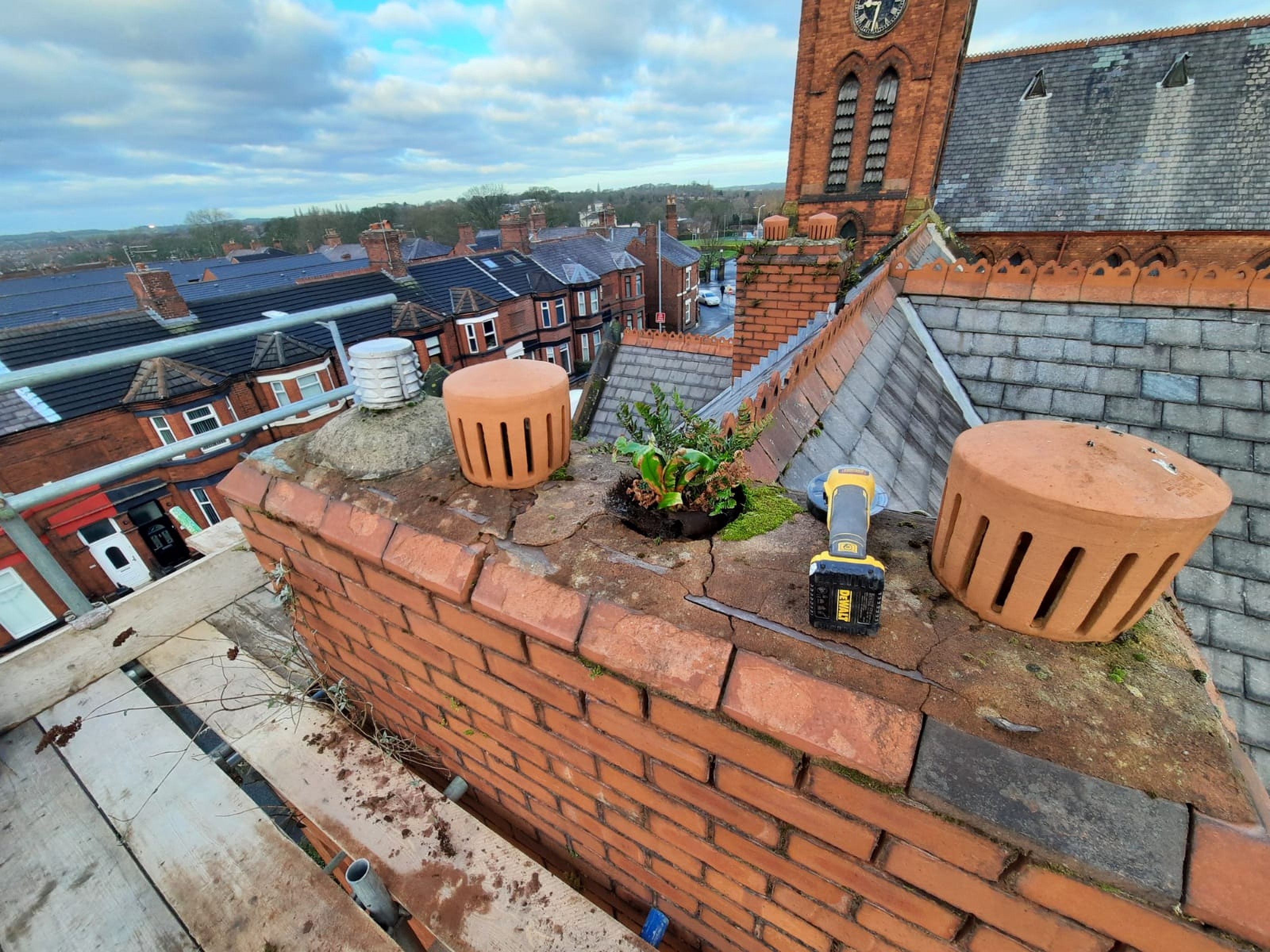 A brick chimney under repair in St Helens with terracotta chimney pots, DeWalt power tool, and scaffolding against a backdrop of a church tower and residential neighbourhood